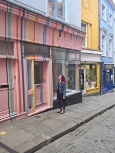 Aimi stands outside the pink colourful shop building, looking up at the window.