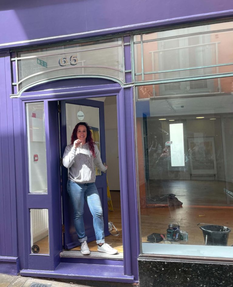 Aimi stands in the doorway of her new shop in blue jeans and a white top. She is holding the key.