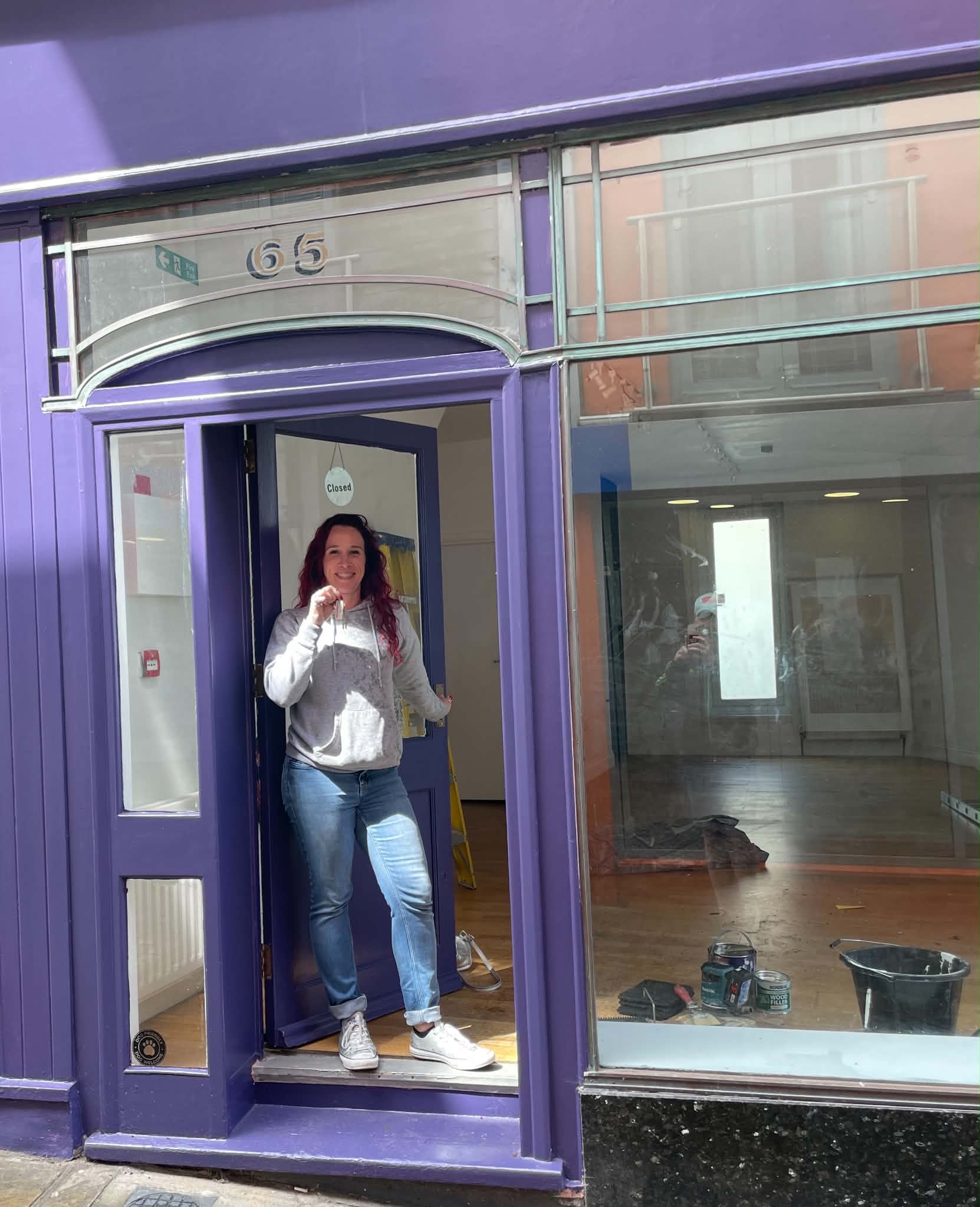 Aimi stands in the doorway of her new shop in blue jeans and a white top. She is holding the key.